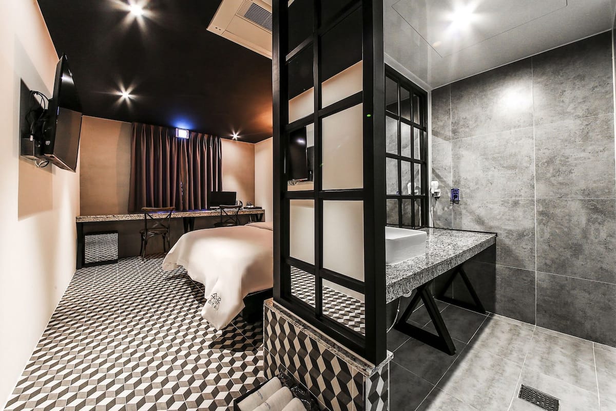 A modern hotel room with a geometric patterned floor, a large bed, a desk with a computer, a bathroom with a granite countertop, and a large mirror.