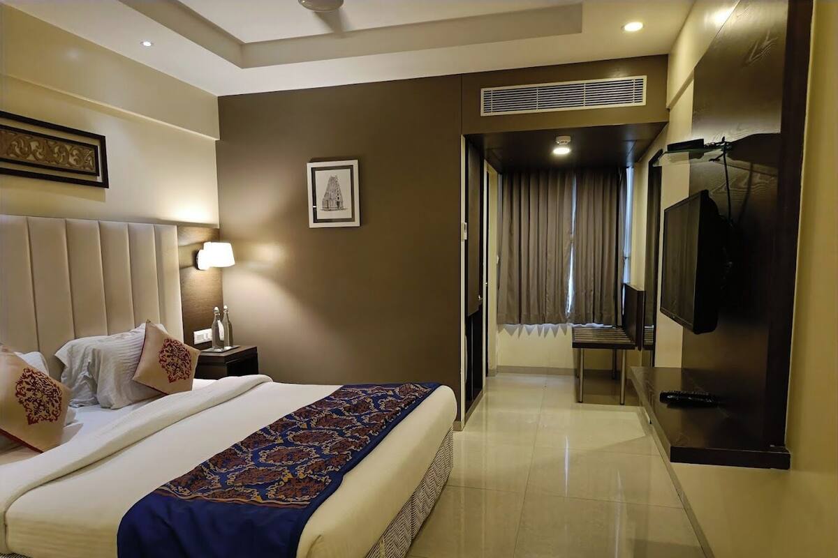 A hotel room with a large bed, a TV, a desk, and a bathroom visible through a glass door.