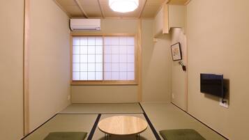 A room with tatami flooring, a low table, two cushions, a window with shoji screens, an air conditioner, and a mounted TV.