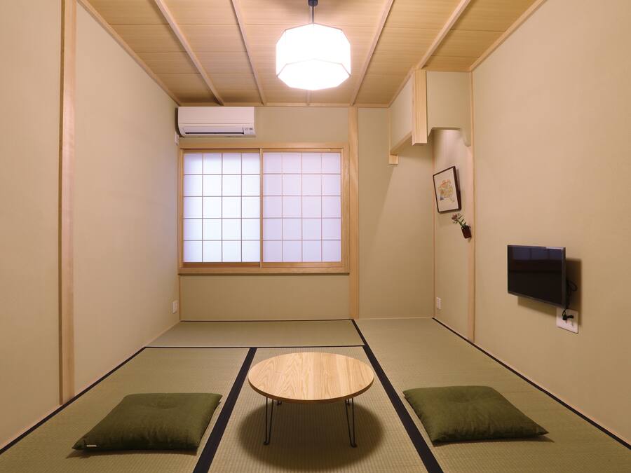 A room with tatami flooring, a low table, two cushions, a window with shoji screens, an air conditioner, and a mounted TV.