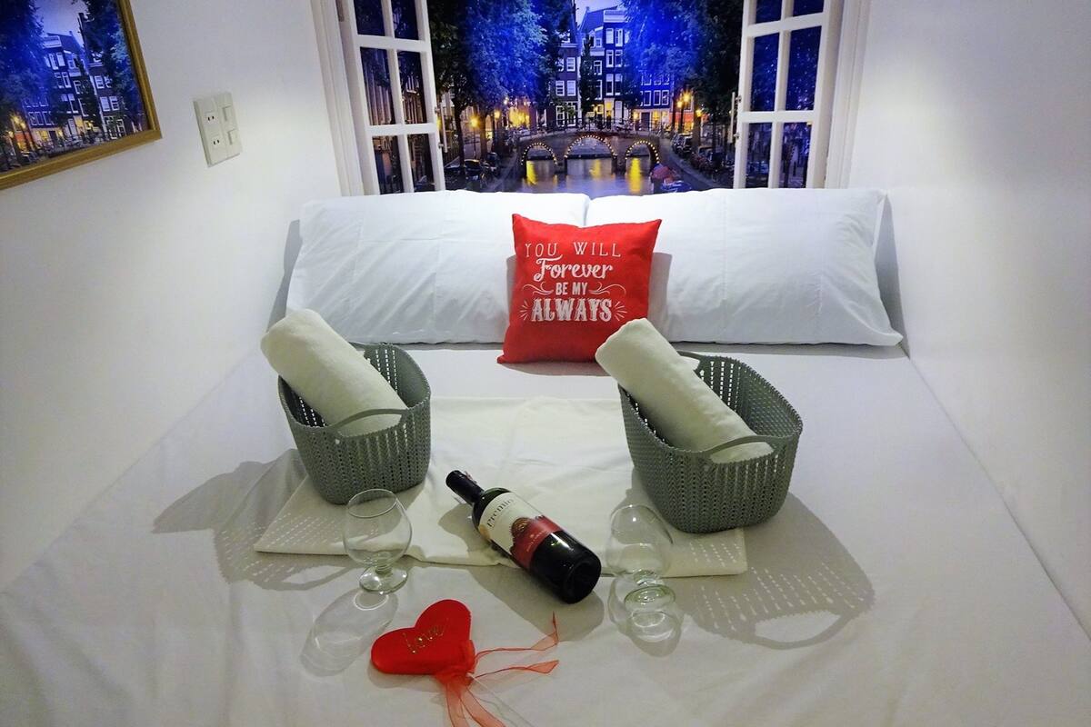 A neatly made bed with white linens, two green baskets, a bottle of wine, a glass, and a red heart-shaped decoration.
