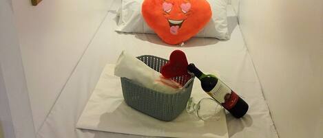 A bed with a view of a coastal landscape, a heart-shaped pillow, a basket with a bottle, and a framed picture on the wall.