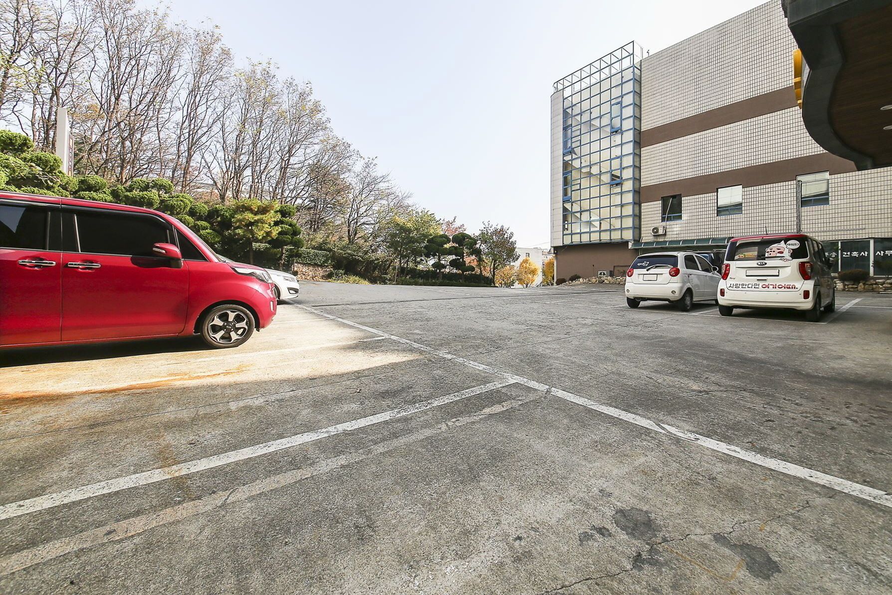 Un estacionamiento con varios autos, incluyendo un auto rojo y un auto blanco con texto coreano. Se observa un edificio moderno con fachada de vidrio al fondo.