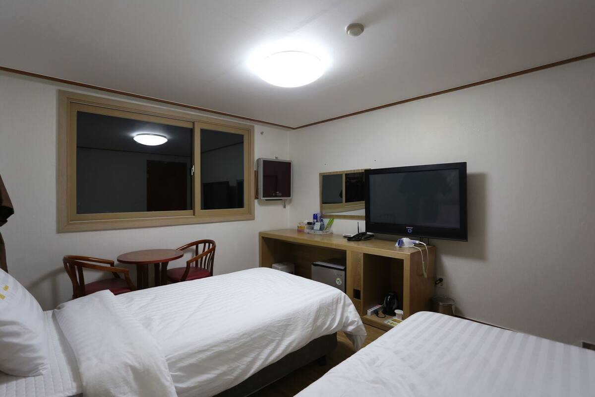 A hotel room with two beds, a desk with a TV, a chair, a small table, and a window.