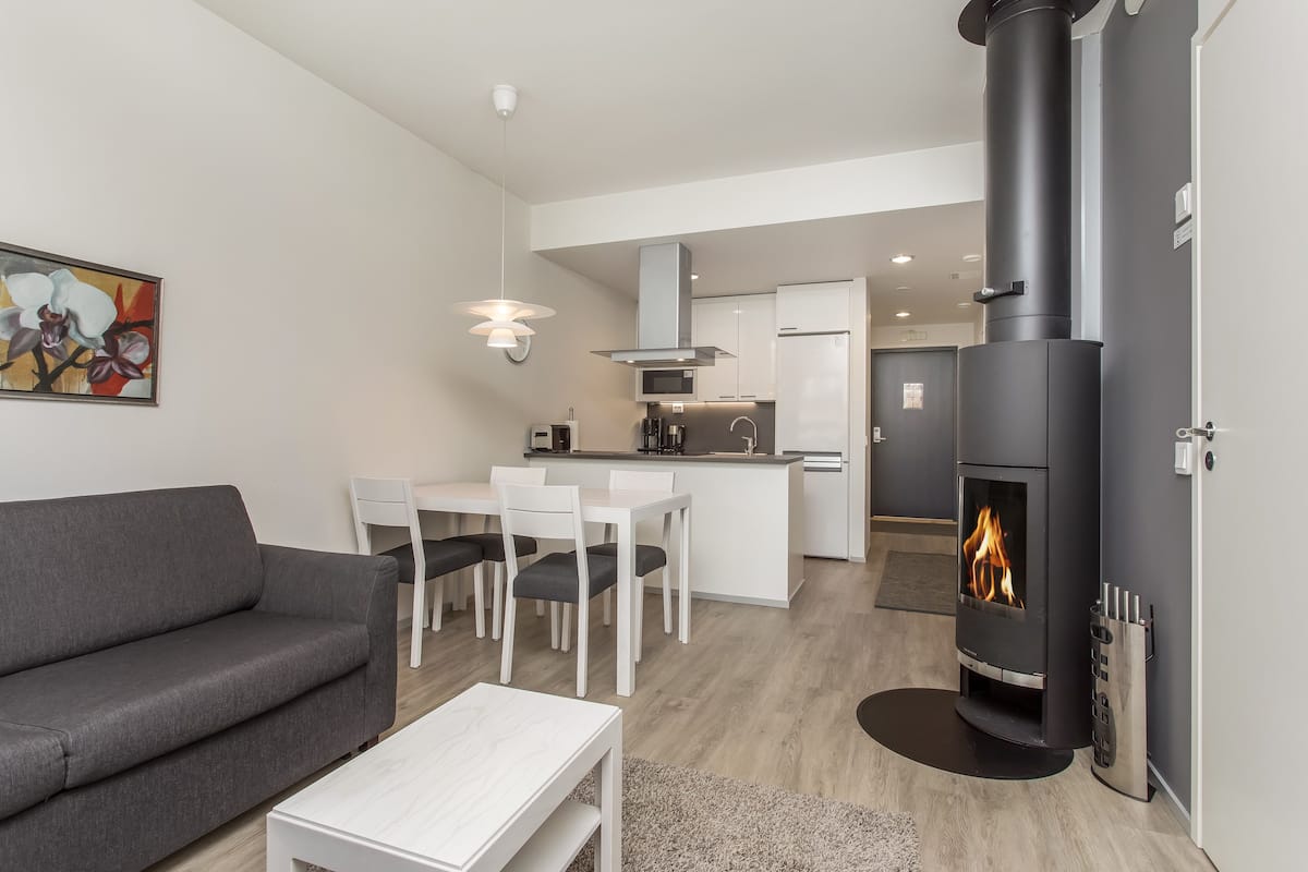 Un salon moderne avec un canapé gris, une table à manger et des chaises blanches, une cuisine équipée d’une cuisinière et un tableau accroché au mur.