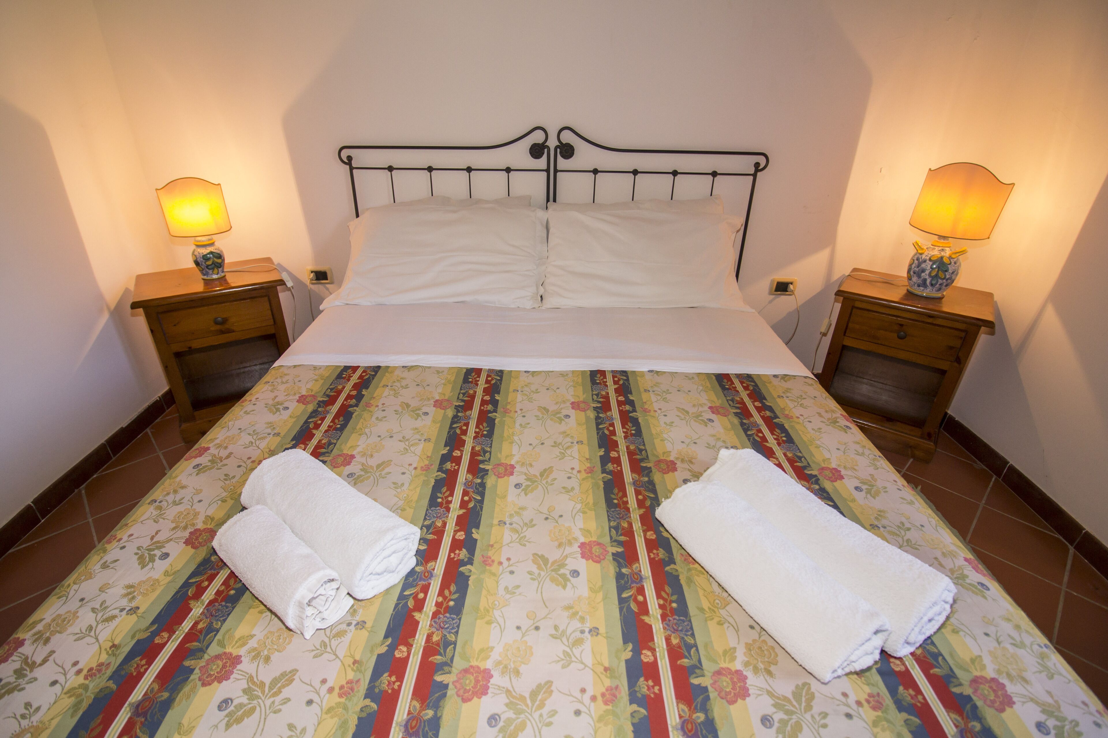 A neatly made bed with a floral-patterned bedspread, two bedside tables with lamps, and two rolled towels on the bed.