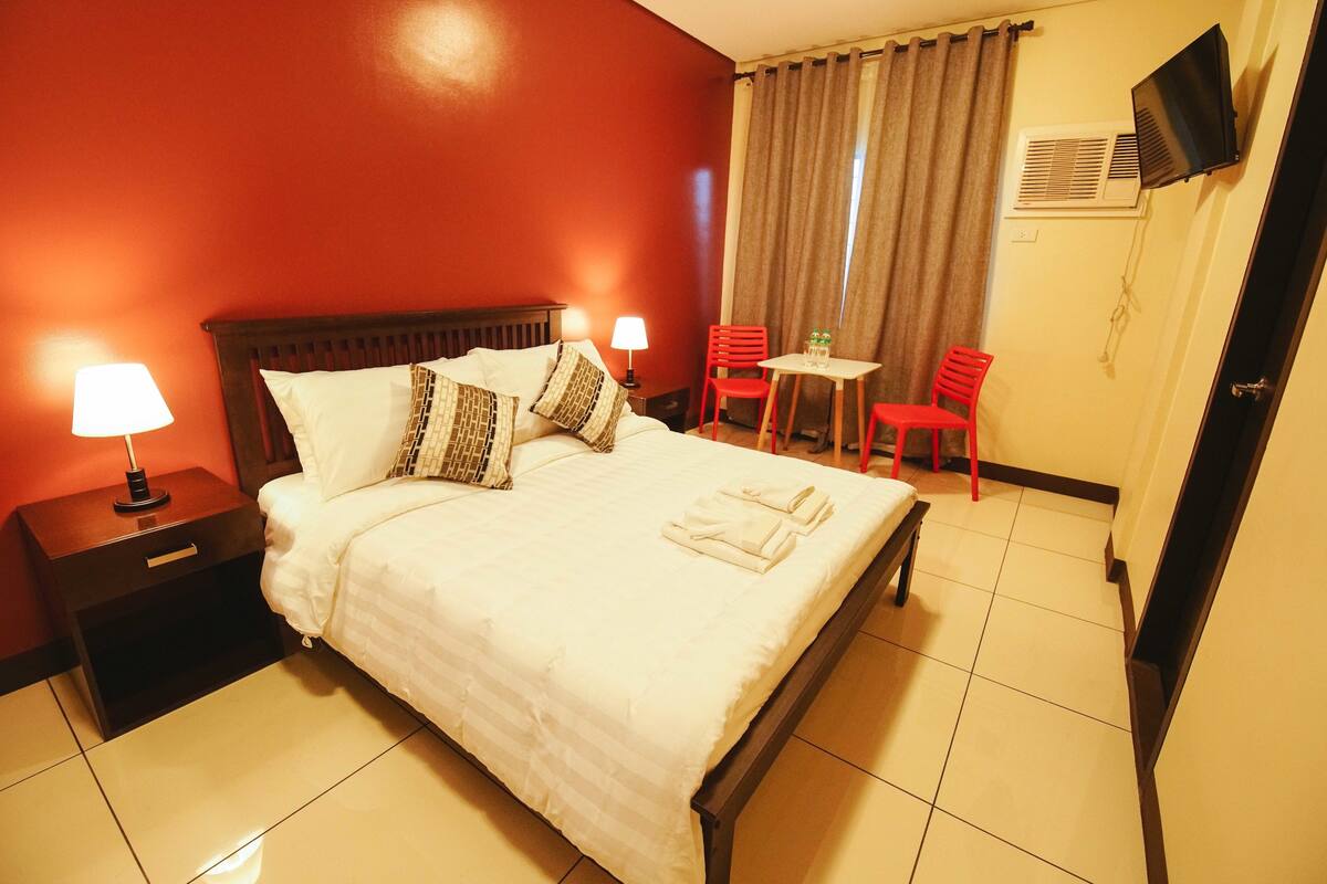 A hotel room with a bed, two bedside tables with lamps, a TV mounted on the wall, a small table with two chairs, and a door.