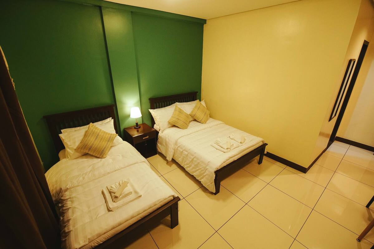 A hotel room with two beds, a nightstand with a lamp, and a green accent wall.