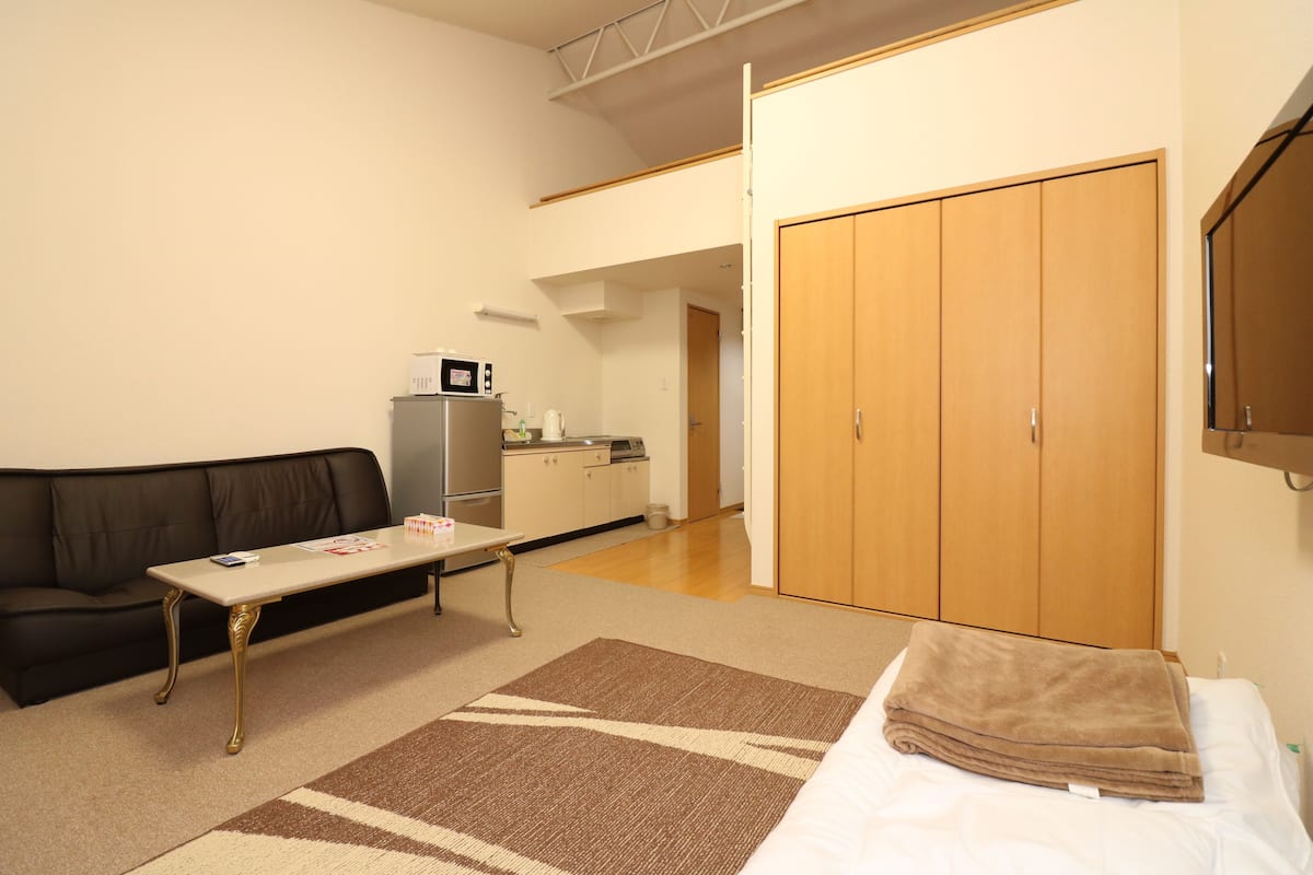 Un espacio habitable pequeño con un sofá, una mesita, una cama con manta, kitchenette y un armario.