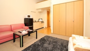 A compact living space with a pink sofa, a small coffee table, a black rug, a kitchen area, and a wardrobe. - BK Hotel & Weekly (Wakayama)