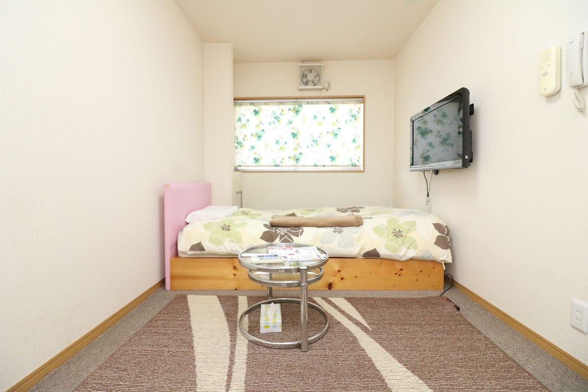 Una habitación pequeña y luminosa con una cama, una mesita, un televisor montado en la pared y una ventana con un estampado floral.