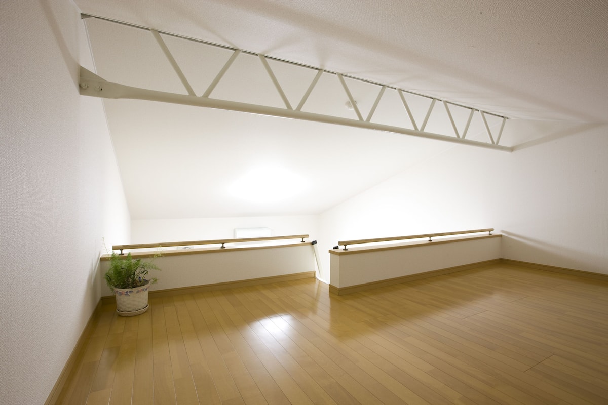 Una habitación con suelo de madera, paredes blancas y techo inclinado. Hay una planta en maceta en la esquina y una ventana con marco de metal.