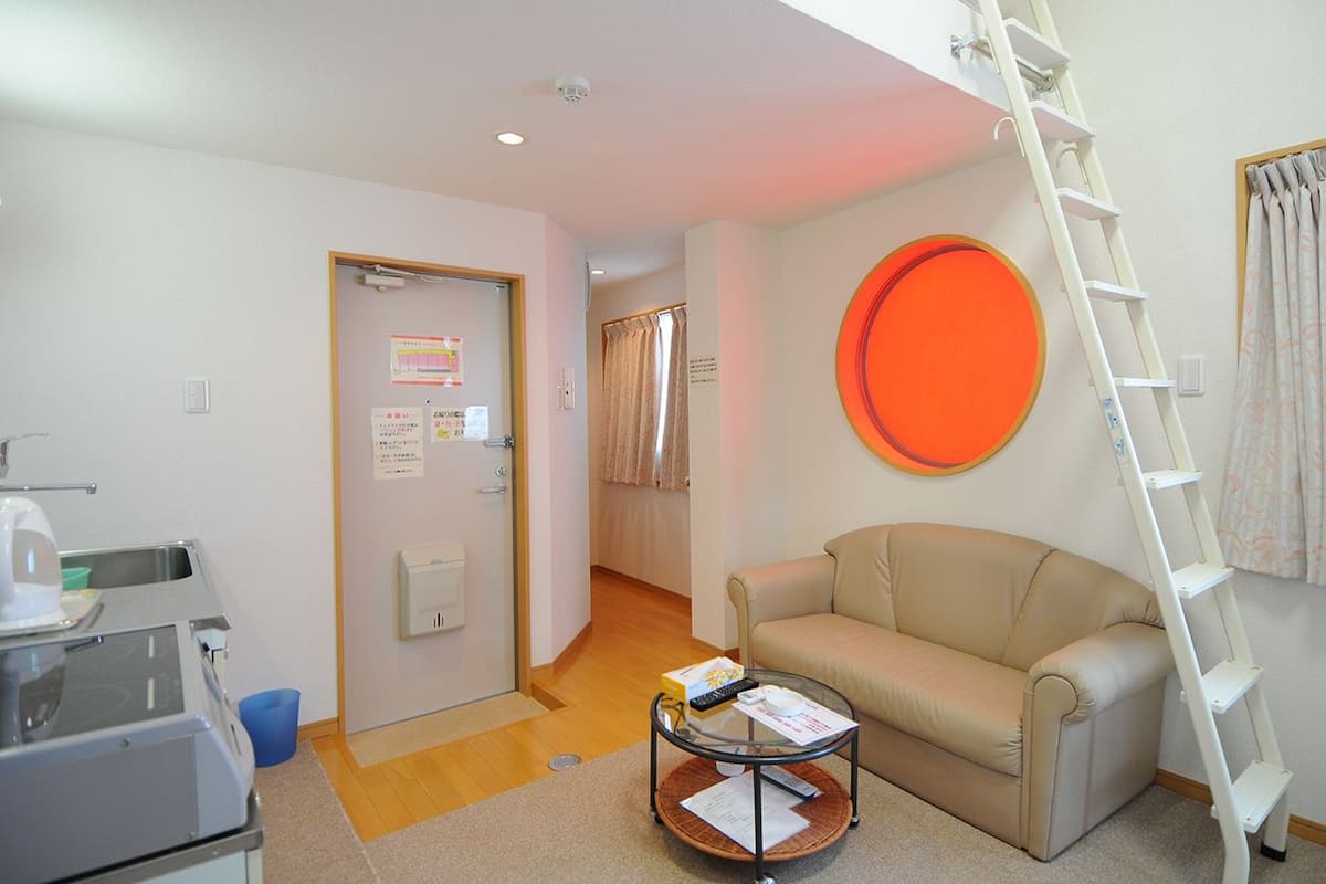 Una sala de estar con un sofá, una mesa de centro, una kitchenette, una puerta con un aviso, una escalera y un gran círculo naranja en la pared.