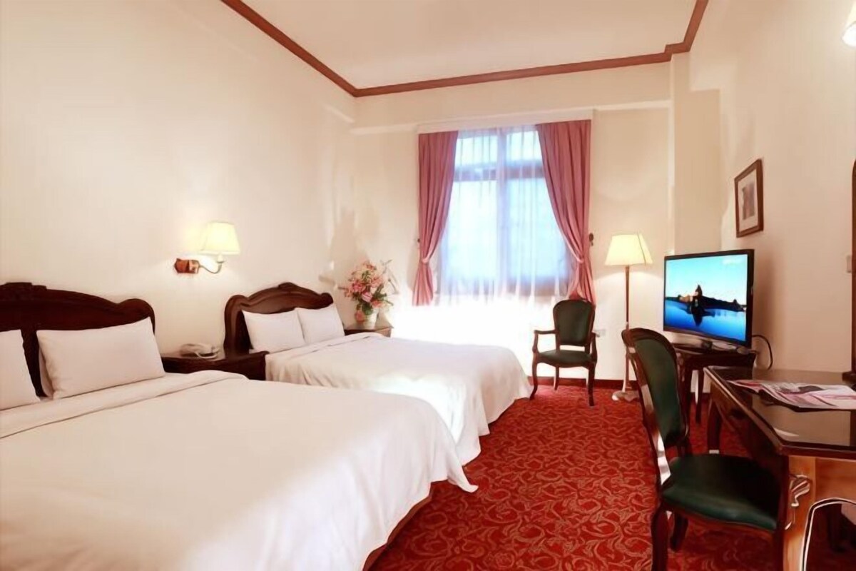 Une chambre d’hôtel avec deux lits, un bureau, une chaise, une télévision et une fenêtre avec des rideaux.