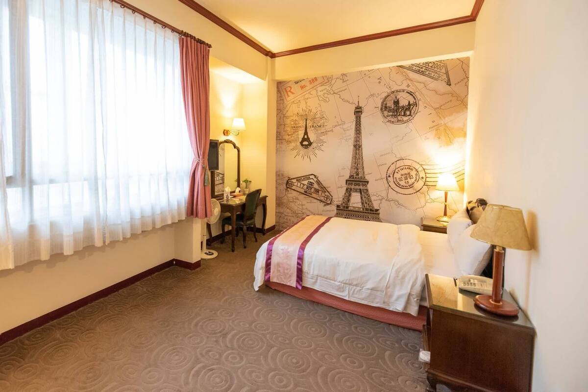 Habitación de hotel con cama, escritorio, silla, mapa en la pared y ventana con cortinas.