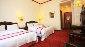 Quarto de hotel com duas camas, uma escrivaninha e uma cadeira. Há luminárias fixadas na parede e um quadro.