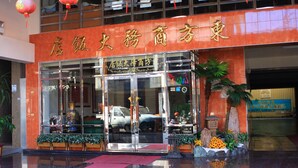 Entrada de hotel com fachada de vidro, tapete vermelho e uma placa com caracteres chineses.