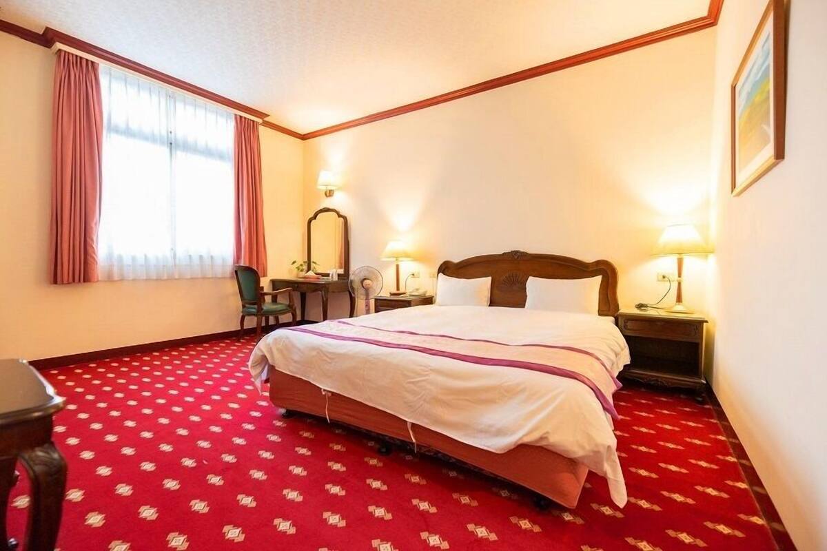 Una habitación de hotel con una cama grande, dos lámparas de noche, un escritorio con silla y una ventana con cortinas.