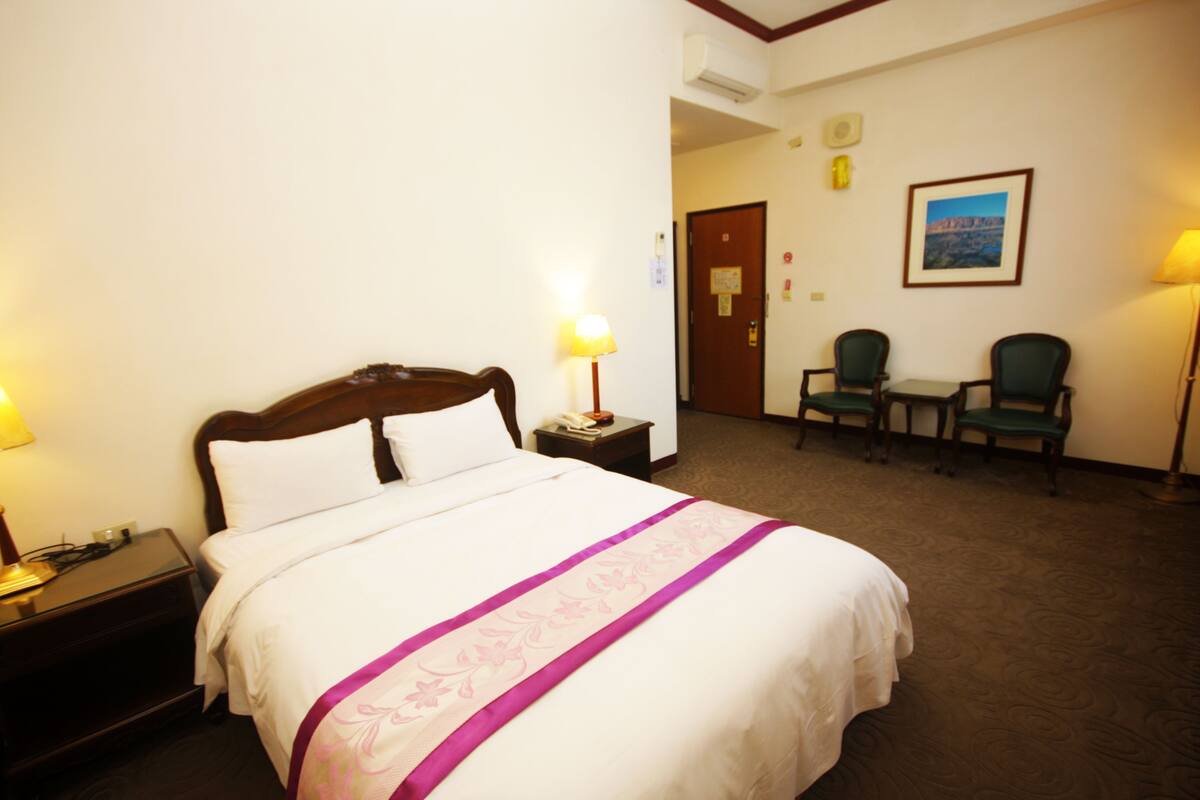 Una habitación de hotel con cama, mesita de noche, lámpara, silla, escritorio y un cuadro en la pared.