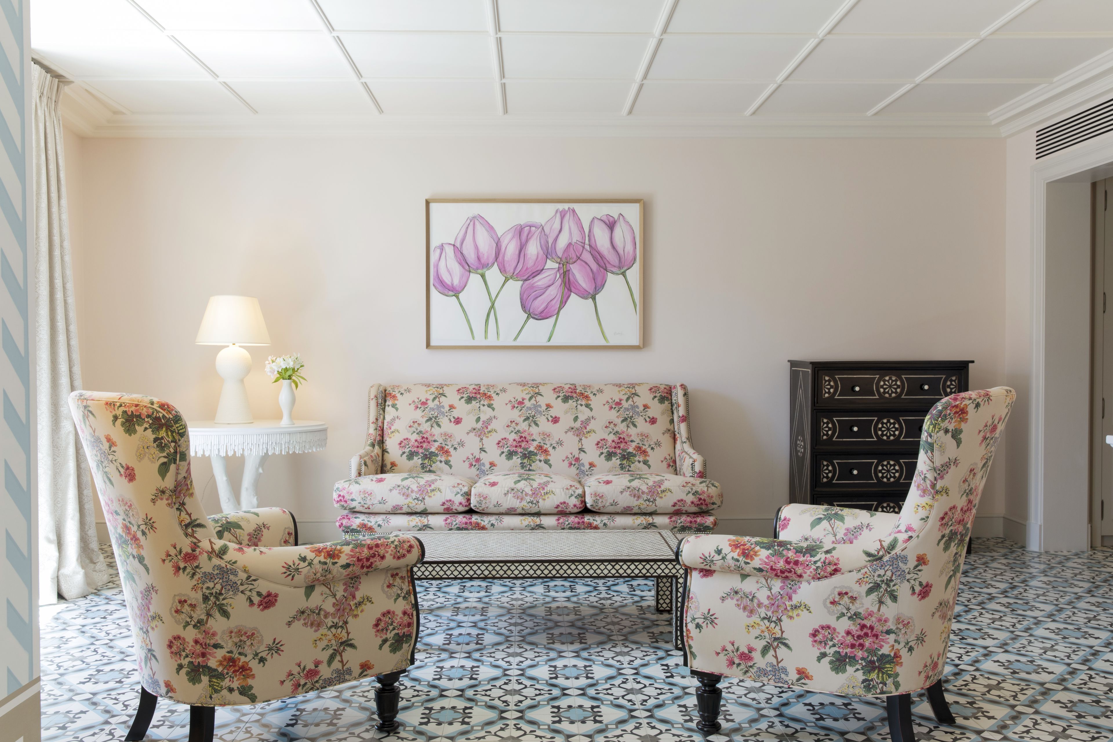 A room with a floral sofa, two armchairs, a patterned floor, a wall-mounted painting, a lamp, and a small table.