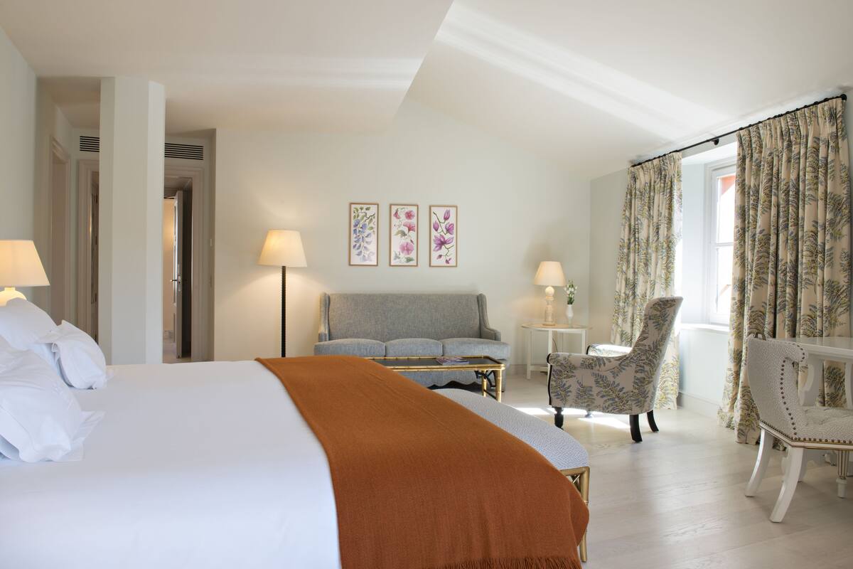 A hotel room with a large bed, a sofa, two armchairs, a small table, and a window with curtains.