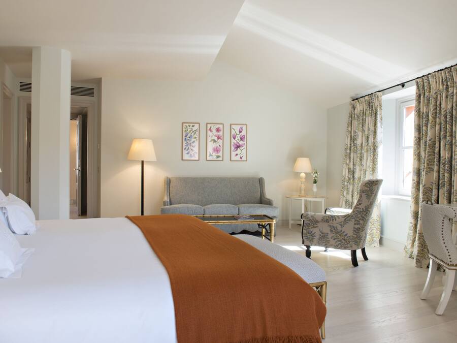 A hotel room with a large bed, a grey sofa, a small table, two chairs, and floral curtains.