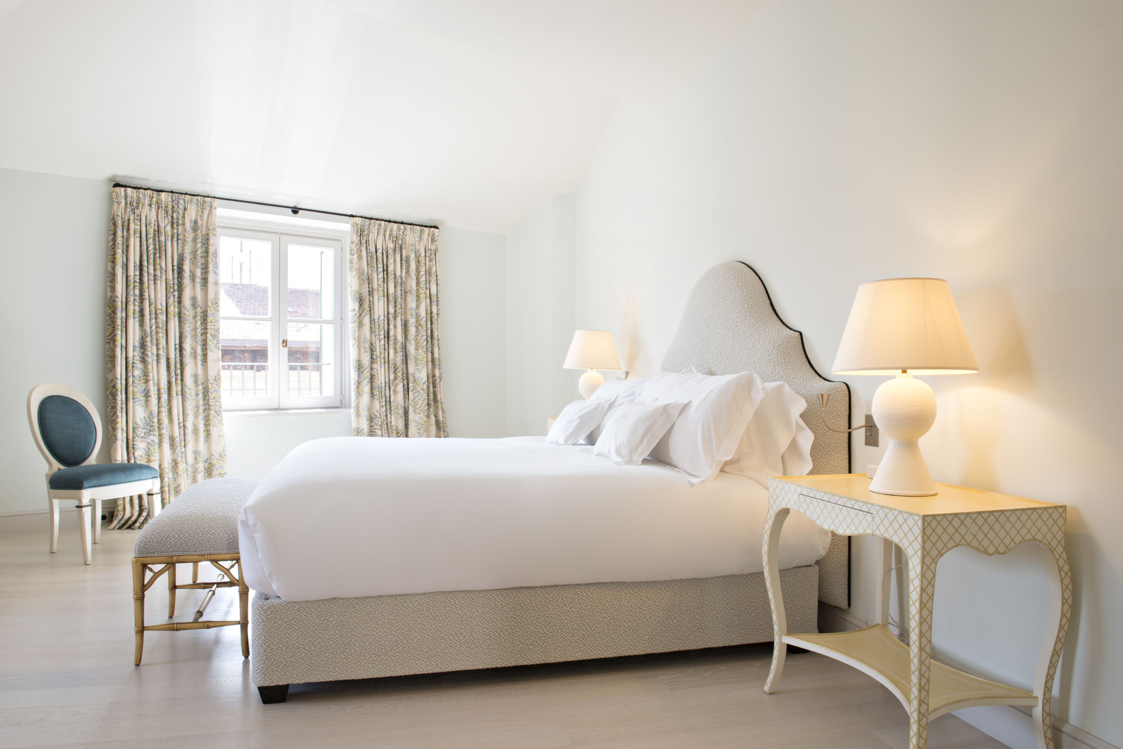 Un dormitorio ordenado con cama, dos lámparas de noche, una silla y una ventana con cortinas.