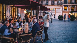 Un café al aire libre con sillas y mesas de mimbre, rodeado de edificios con balcones y grandes ventanales.
