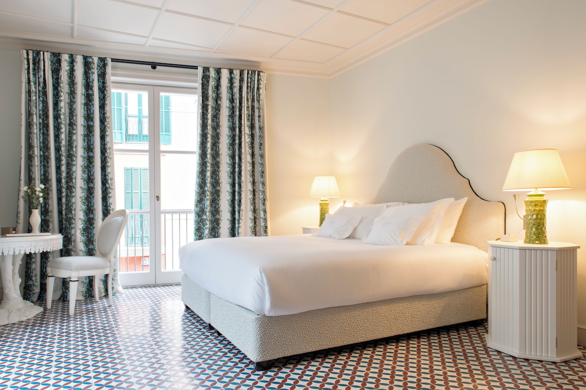 A hotel room with a large bed, two bedside lamps, a chair, a small table, and patterned floor tiles.