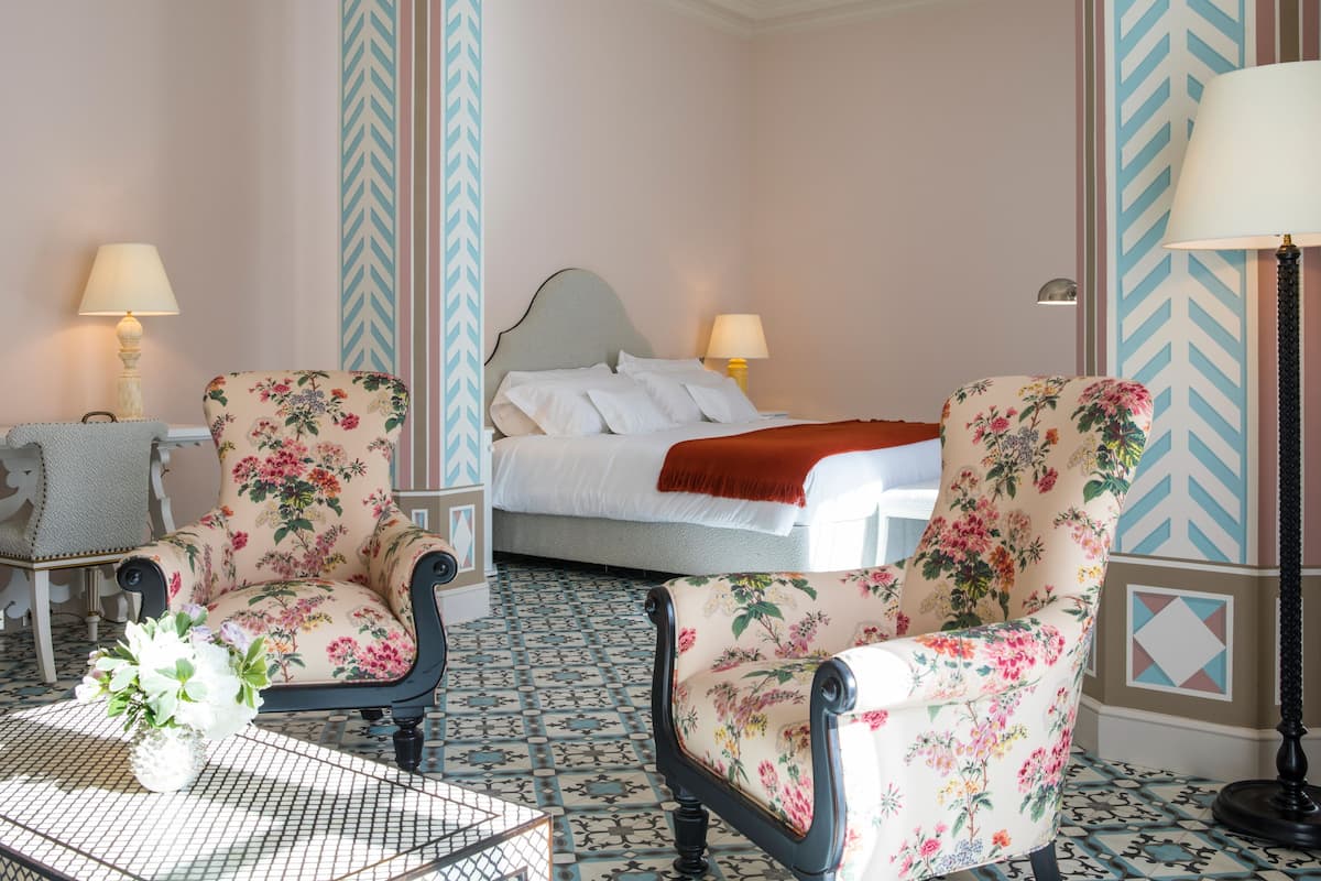 A hotel room with a bed, two armchairs with floral upholstery, a small table with a vase, and patterned floor tiles.
