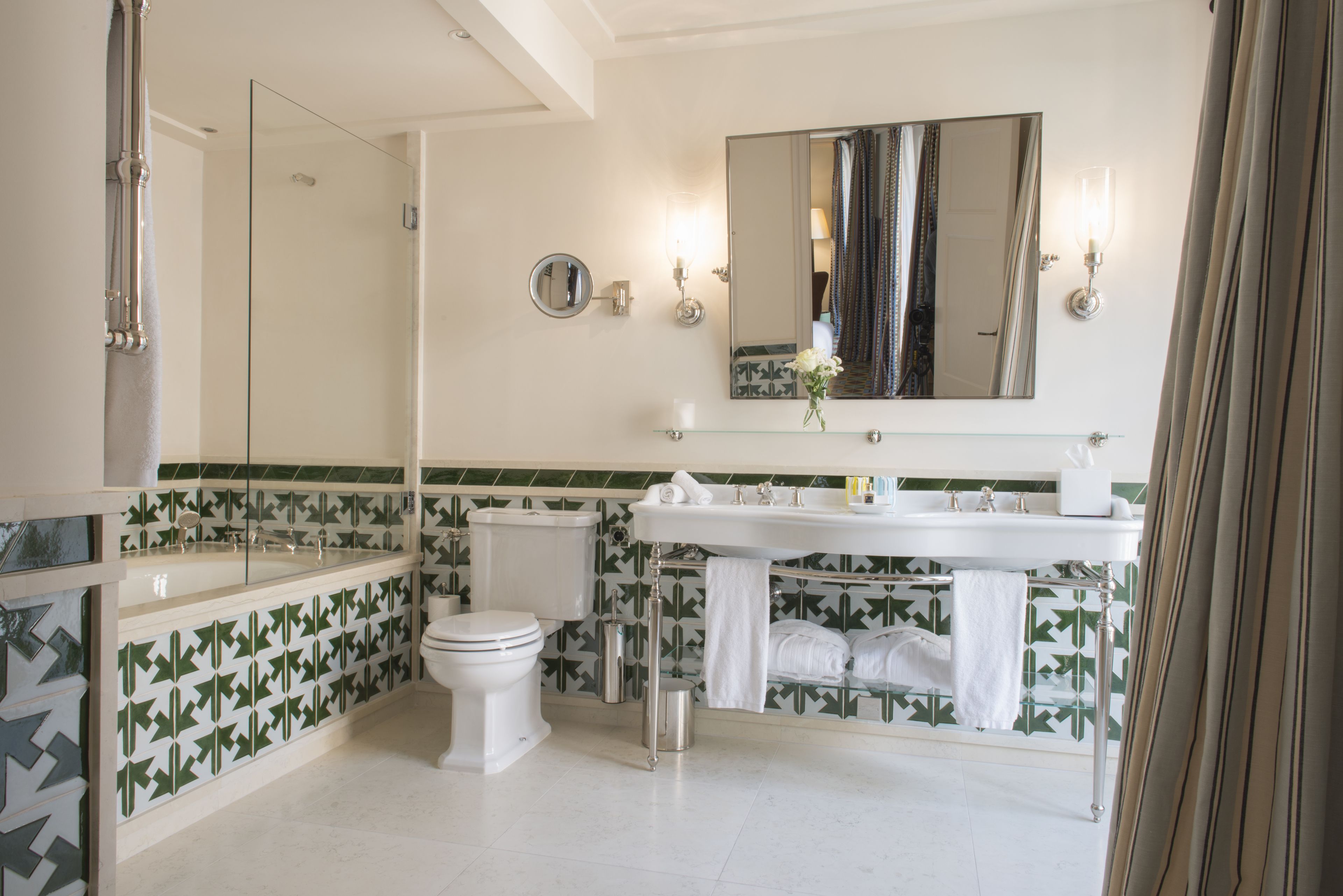 Une salle de bain moderne avec une douche à parois de verre, des toilettes blanches, un lavabo avec un miroir au-dessus et des carreaux verts et blancs à motifs.