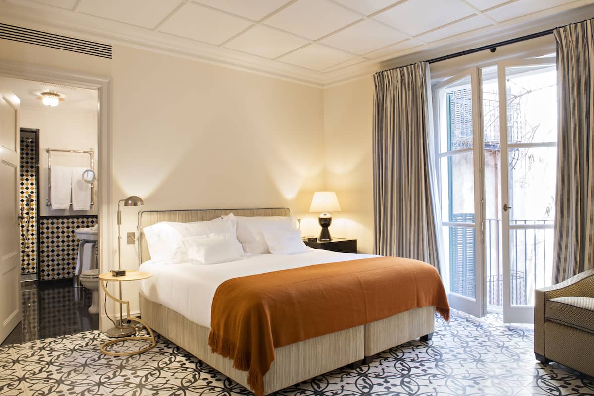 A hotel room with a large bed, a bedside table with a lamp, a bathroom with a toilet and sink, and a view of the outside through a door.