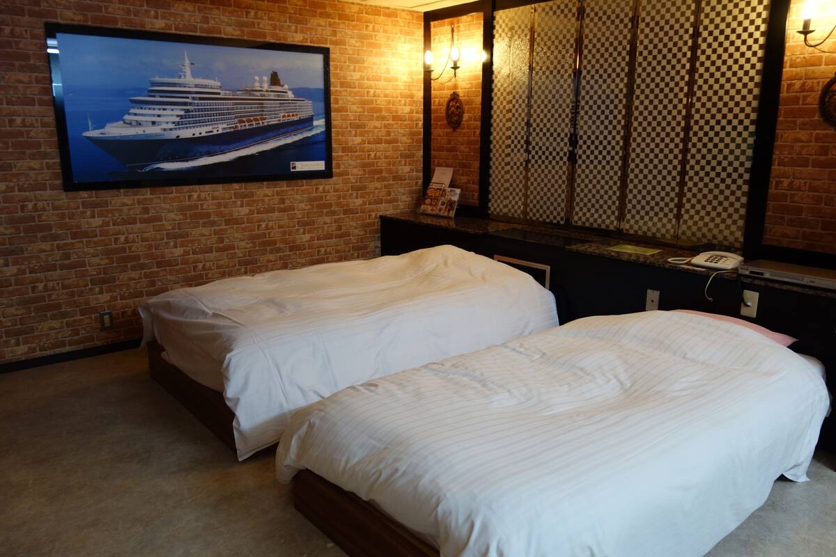 A hotel room with two beds, a large window, a television displaying a cruise ship, and a checkered pattern on the wall.