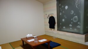 A room with tatami flooring, a wooden table with a tissue box and papers, a blue cushion, and a wall with a decorative hanging. - Sea Queen Kainan (Kainan)