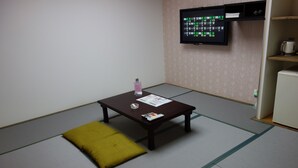 A room with tatami mat flooring, a small wooden table with a magazine, a bottle, and a pillow. A television is mounted on the wall, and a refrigerator is visible in the background. - Sea Queen Kainan (Kainan)