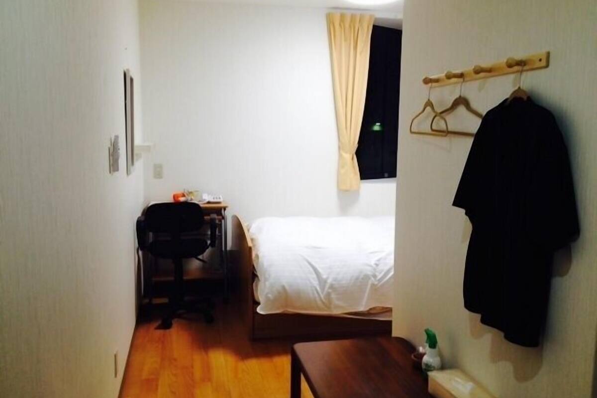 A small, simple room with a bed, a desk, and a chair. There is a coat rack with a jacket hanging on it.