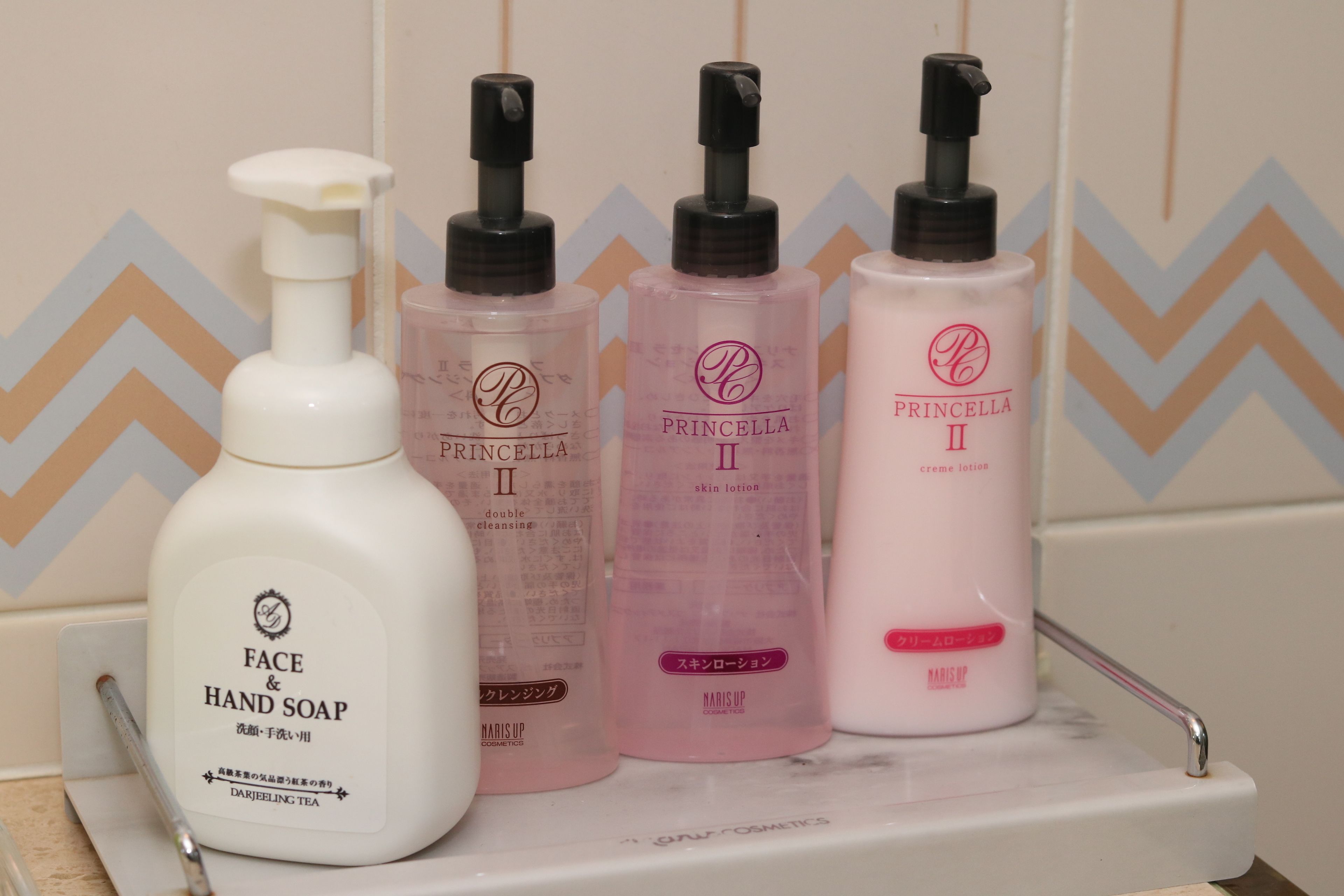 Uma prateleira no banheiro com diversos produtos de higiene, incluindo uma garrafa de sabonete líquido para rosto e mãos, duas garrafas de loção Princess II para a pele e uma garrafa rosa de creme hidratante Princess II.