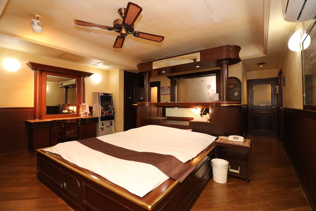 A hotel room with a large bed, a wooden dresser, a vanity with a mirror, a ceiling fan, and a small trash bin.