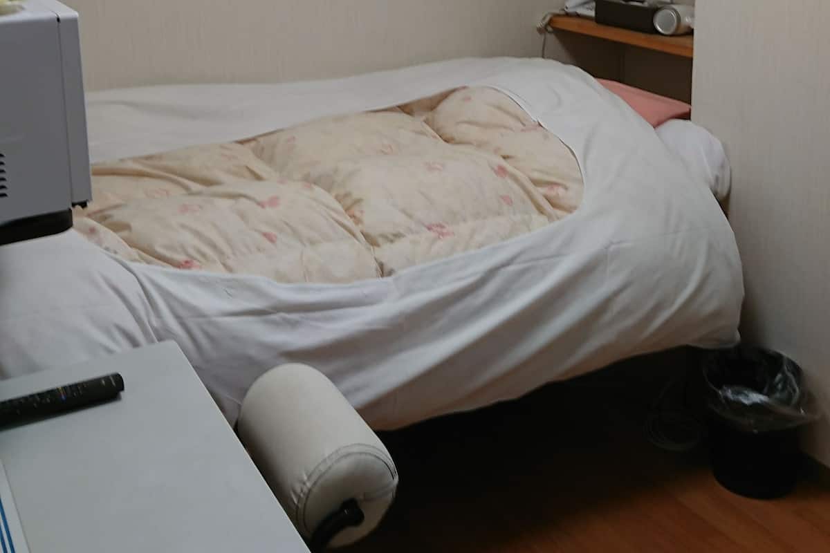 A small, simple room with a bed, a desk with a computer, a chair, a small table with a phone and a radio, and a trash bin.