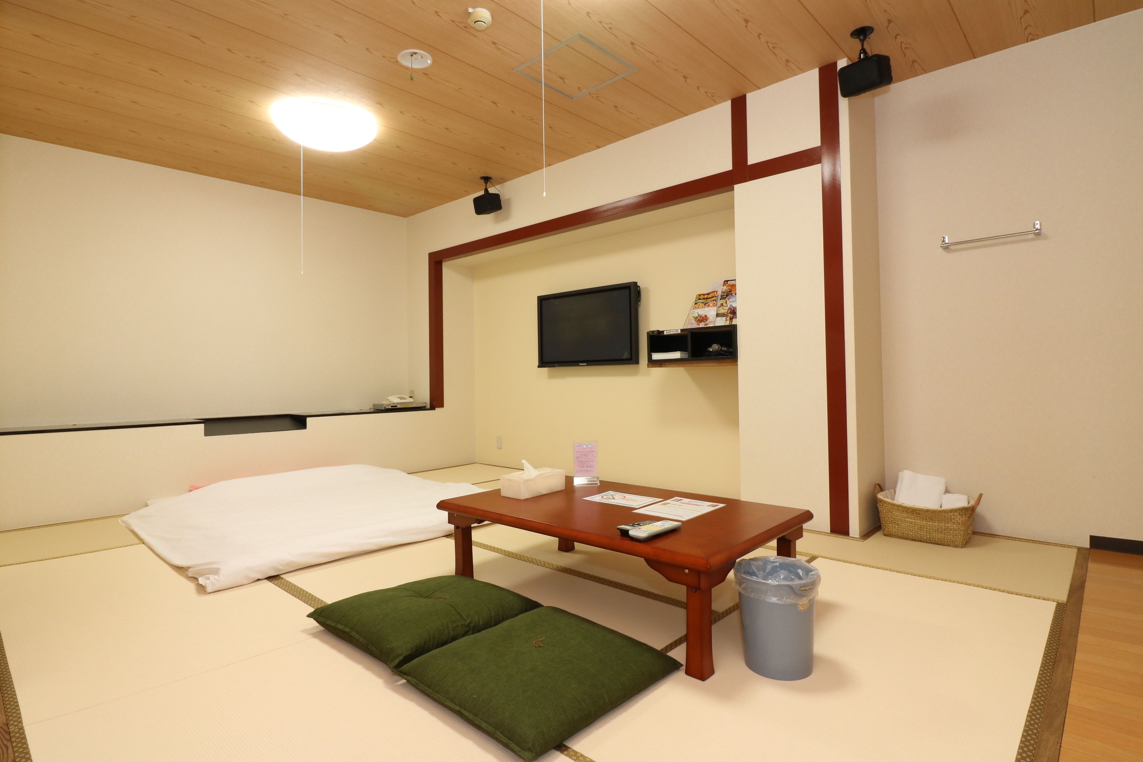 A room with tatami flooring, a futon bed, a wooden table with a remote control, a trash bin, and a television mounted on the wall.