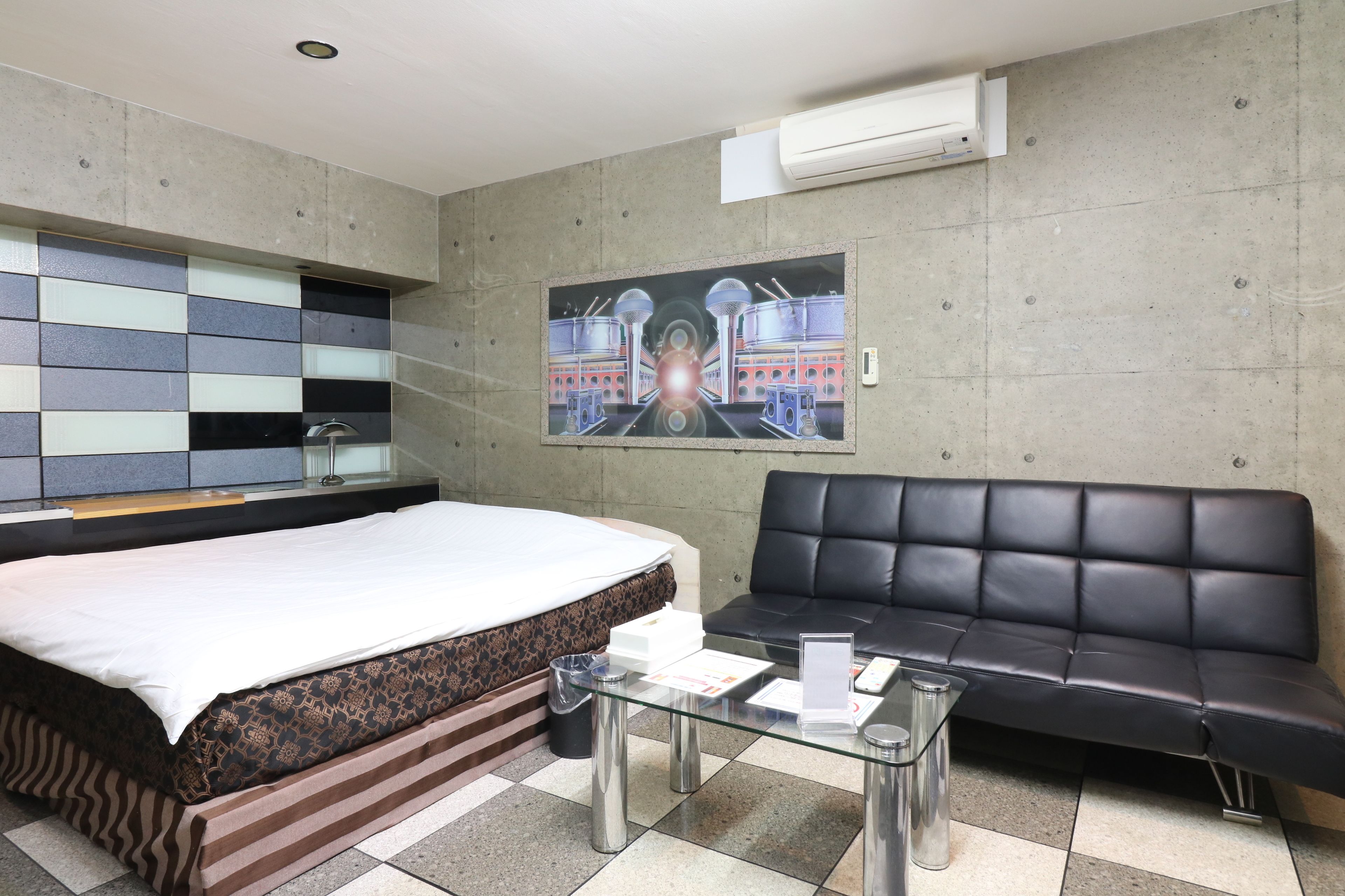 A hotel room with a bed, a black leather sofa, a glass coffee table, and a painting on the wall.