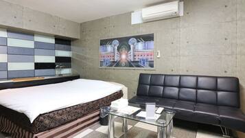 A hotel room with a bed, a black leather sofa, a glass coffee table, and a painting on the wall.