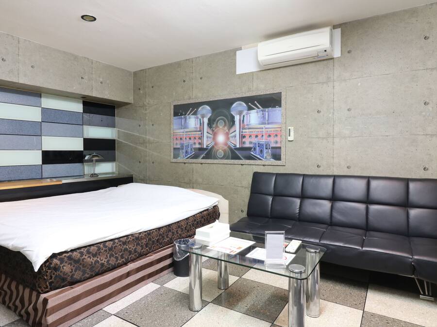 A hotel room with a bed, a black leather sofa, a glass coffee table, and a painting on the wall.