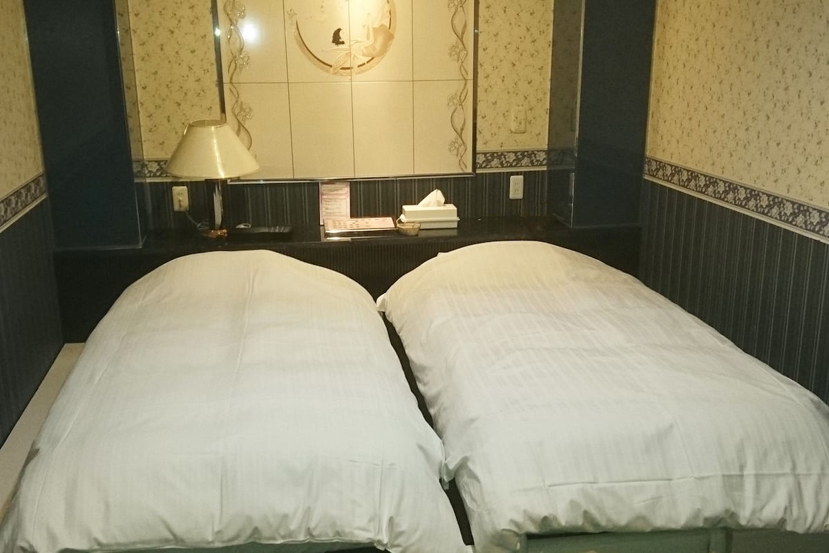A double bed with white linens in a hotel room with a marble floor and a wall-mounted light fixture.