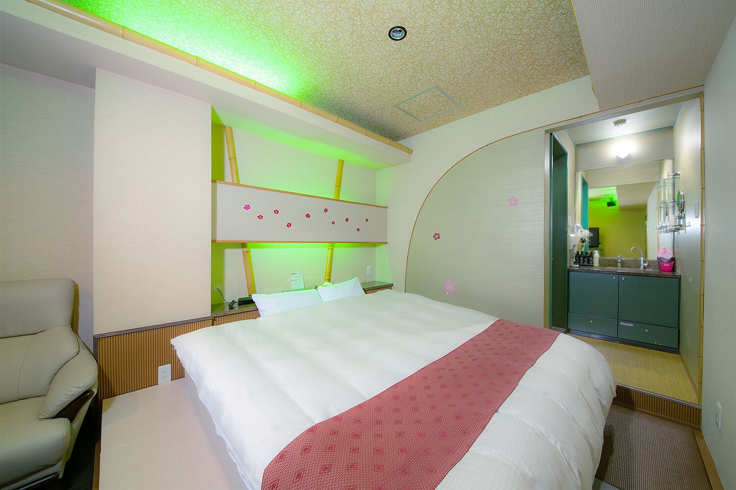 A hotel room with a bed, a chair, a vanity with a mirror, and a green-lit wall.