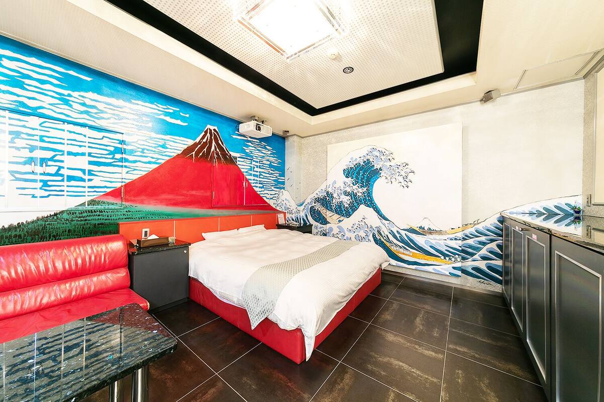 A hotel room with a bed, a red sofa, a desk, and a large mural of a wave and mountain.