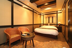 Een hotelkamer met een groot bed, twee rieten stoelen, een klein glazen tafeltje en een spiegel aan de muur.