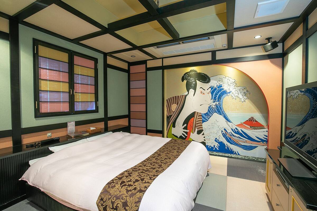 A hotel room with a bed, a TV, and a wall with a Japanese wave painting.