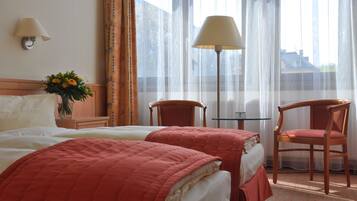 A hotel room with two beds, a table, two chairs, a lamp, and a window with sheer curtains.