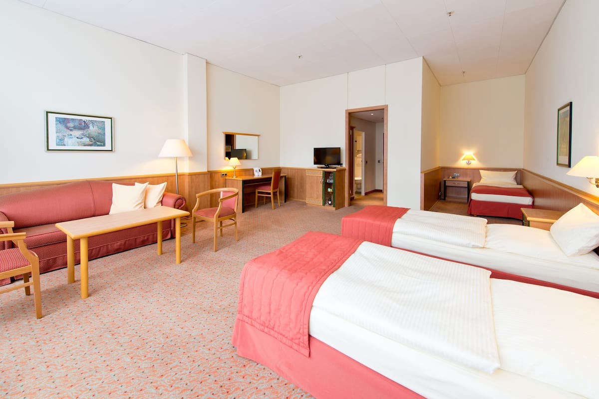 A hotel room with a sofa, two single beds, a desk with a chair, a TV, and a lamp.